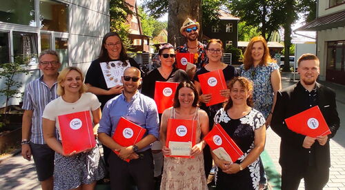Klassenfoto. Gruppenfoto draußen unter einem Baum. 8 Menschen halten fröhlich lächelnd einen rote AWO A4 Mappen mit Gummiband vor sich. Zwei Frauen und ein Mann halten nichts in den Händen.