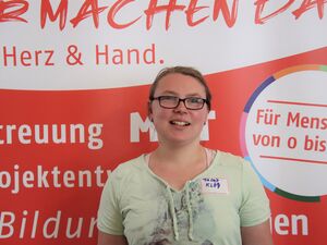 Junge Frau vor einem AWO Aufsteller. Sie trägt eine Brille mit dunklem Rahmen, braune Haare, ei mint-grünes T-Shirt.