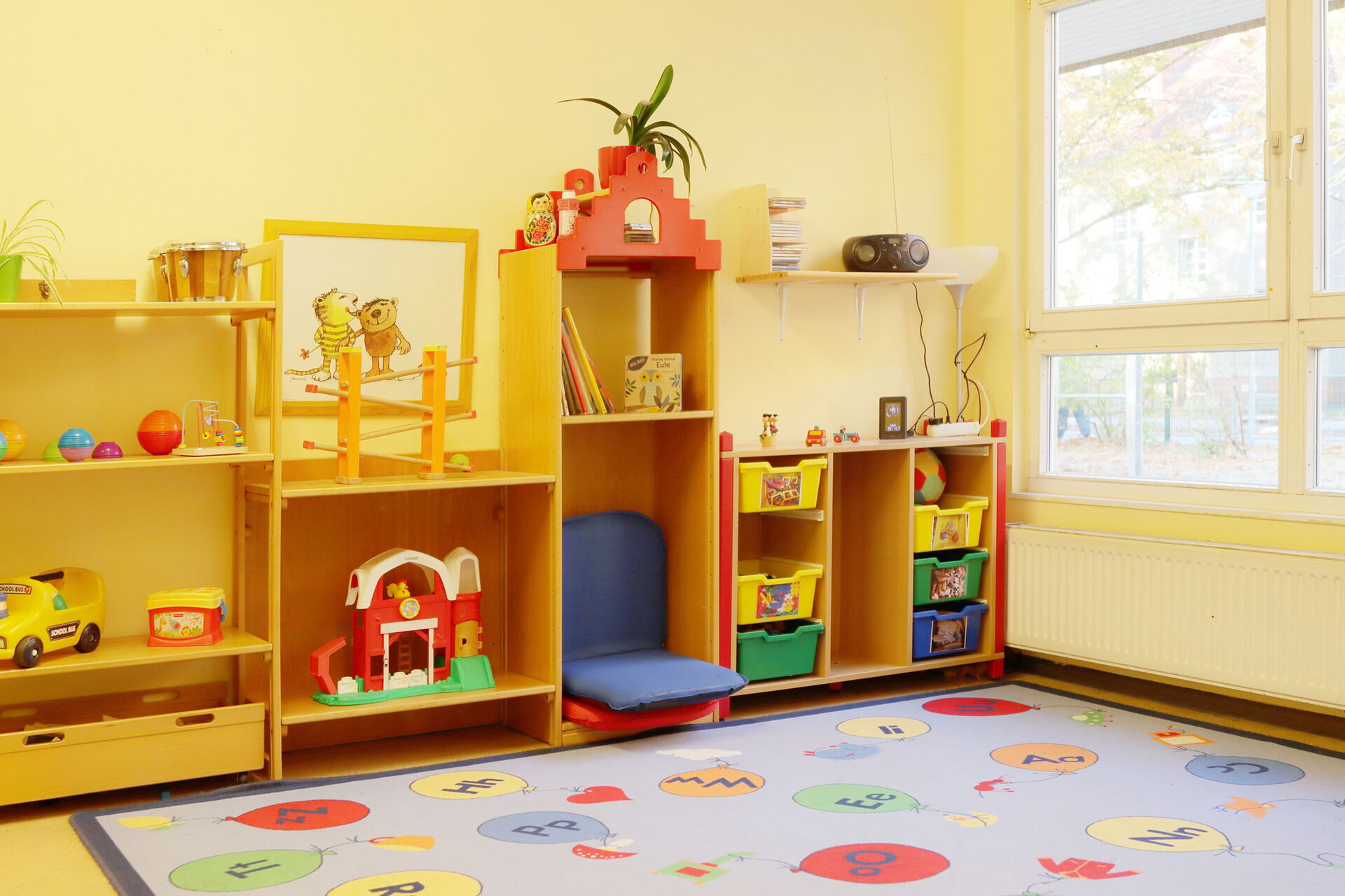 Heller Raum, Regal mit Spielzeug steht an einer gelben Wand, davor ein Teppich mit bunten Buchstaben-Luftballons.