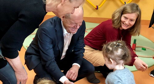 Ein kleines Mädchen tapst auf einen alten Mann mit Glatze im dunklen Anzug und weißem Hemd darunter zu. Der Mann streckt ihr die Hand entgegen. Eine Frau beugt sich vor. Eine Frau mit Kind auf dem Schoß sitzt auf dem Fußboden. Eine weitere Frau hockt nebe