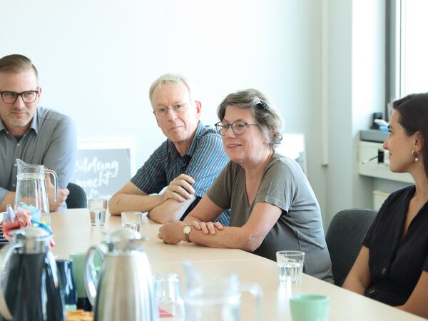 2 Männer und zwei Frauen sitzen am Tisch. Auf dem Tisch Kaffeekannen, Wasserkaraffe, Gläser und Tassen.