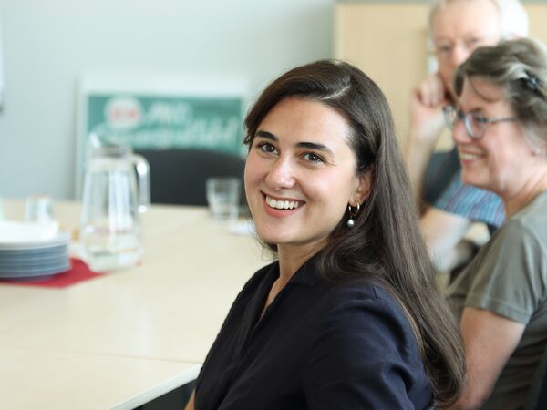 Junge Frau lächelt, hat den Kopf zur Kamera gedreht. Sie sitzt am Tisch. Sie hat lange dunkle Haare und Ohrringe. Zwei weitere Personen sitzen am Tisch sind unscharf dargestellt.