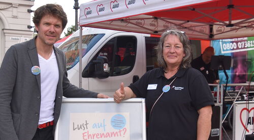 Draußen auf einem Platz. Ein Mann und eine Frau. Die Frau reckt dem Damen nach oben. Zwichen den Beiden ein Schild. Text: Lust auf Ehrenamt?