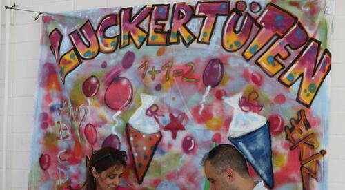 Frau, Kind mit leerer Zuckertüte und mann, vor einem großen Transperen mit Zuckertüten und dem Schriftzug oben drüber.