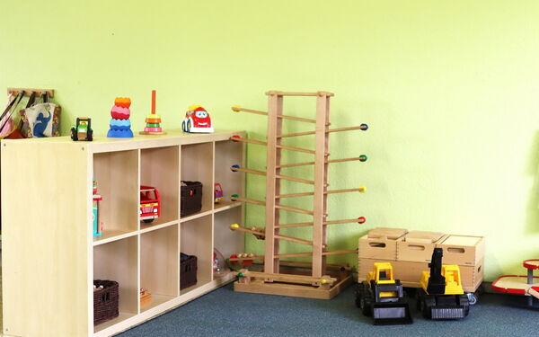 Spielzimmer mit Spielsachen. Ein Regal mit Feuerwehrauto, Trecker, Stapelturm. Auf dem Fußboden stehen eine Holzkugelbahn und zwei gelbe Bagger.
