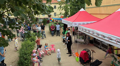 Draufsicht von weiterweg. Kita-Gelände. Rechts zwei rote AWO Pavillons, 4 Kinder sitzen auf kleinen pinken Stühlen. Erwachsene und weitere Kinder stehen herum.