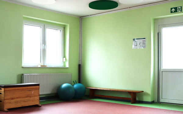 Turnhalle. Grüne Wände. Ein Fenster, eine weiße Tür, mit Notausgang-Schild darüber. Eine Bank steht an der Wand, zwei Pezzi-Bälle und ein Sprungkasten