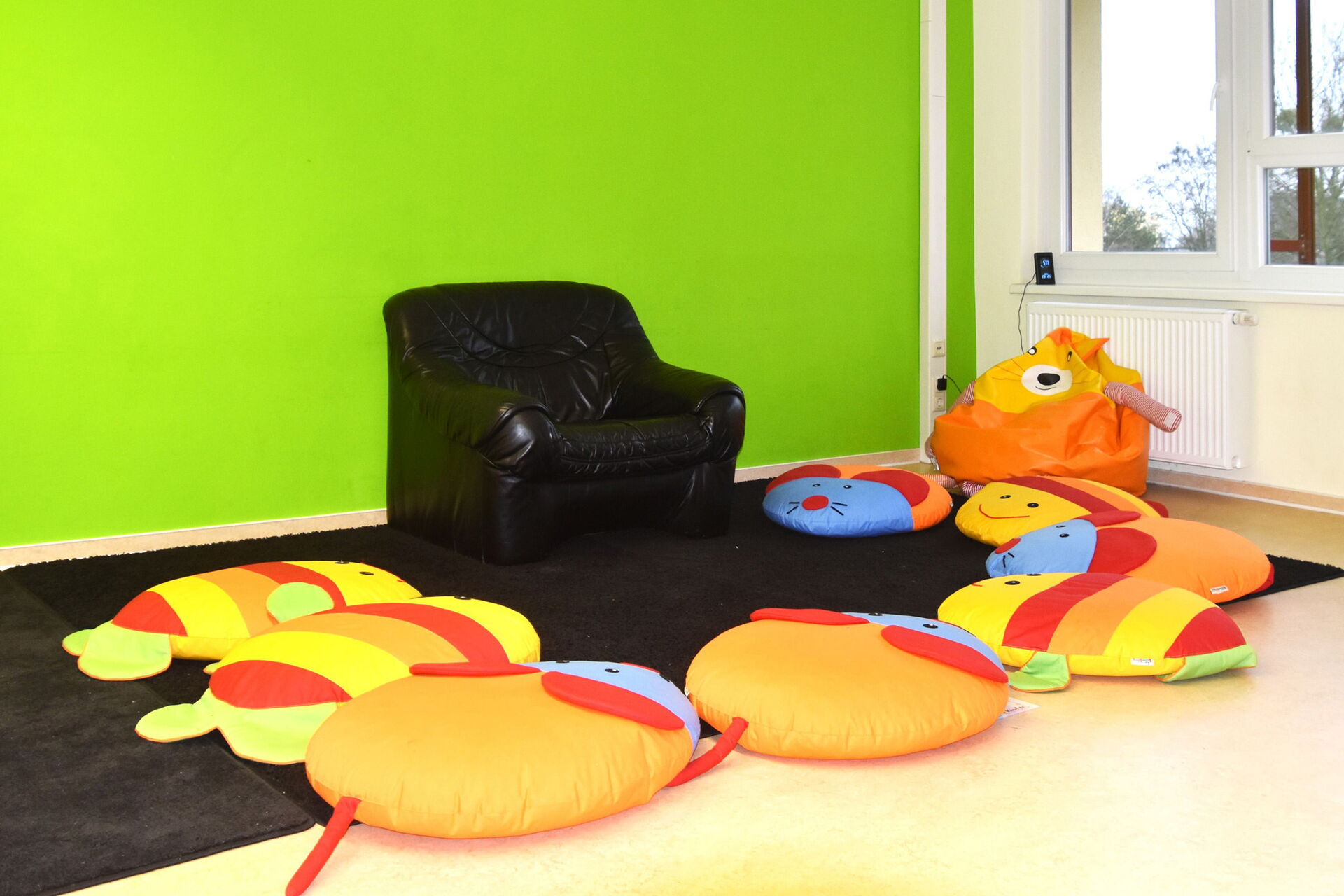 Zimmer mit einer grünen Wand. An der Wand steht ein schwarzer Sessel. Um den Sessel liegenn 8 bunte Sitzkissen, die aussehen wie Mäuse und Fische.