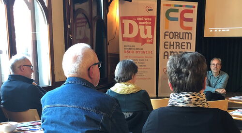 Das Bild zeigt mehrere Menschen im Zuschauerraum, die einem Mann zuhören, der auf der Bühne sitzt.