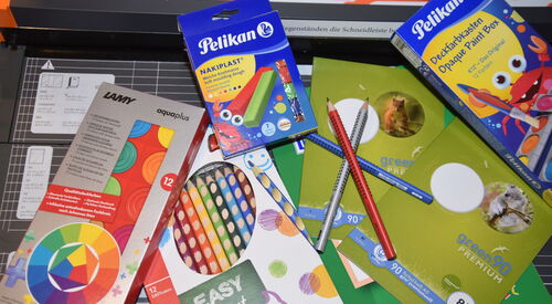 Schulmaterial. Eine Packung mit Buntstiften, Pelikan Knete Nakiplast, Deckfarbkasten von Pelikan, Wasserfarbkasten von LAMY Aquaplus, Schulheft Staufen Recycling green90 Premium