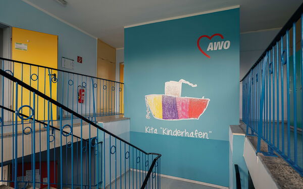 Treppenhaus in Blau. Auf einer Wand das AWO Logo und ein buntes Schiff mit Anker. Unter dem Schiff steht in weißer Schrift Kita "Kinderhafen".