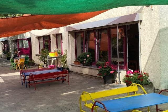 Farbige Bänke und Tische vor einem Gebäude. Bepflanzte Blumenkübel auf einer Fensterbank und zu beiden Seiten einer Terassentür.