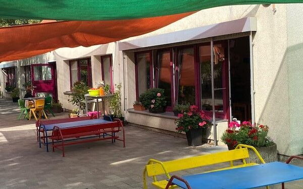 Farbige Bänke und Tische vor einem Gebäude. Bepflanzte Blumenkübel auf einer Fensterbank und zu beiden Seiten einer Terassentür.