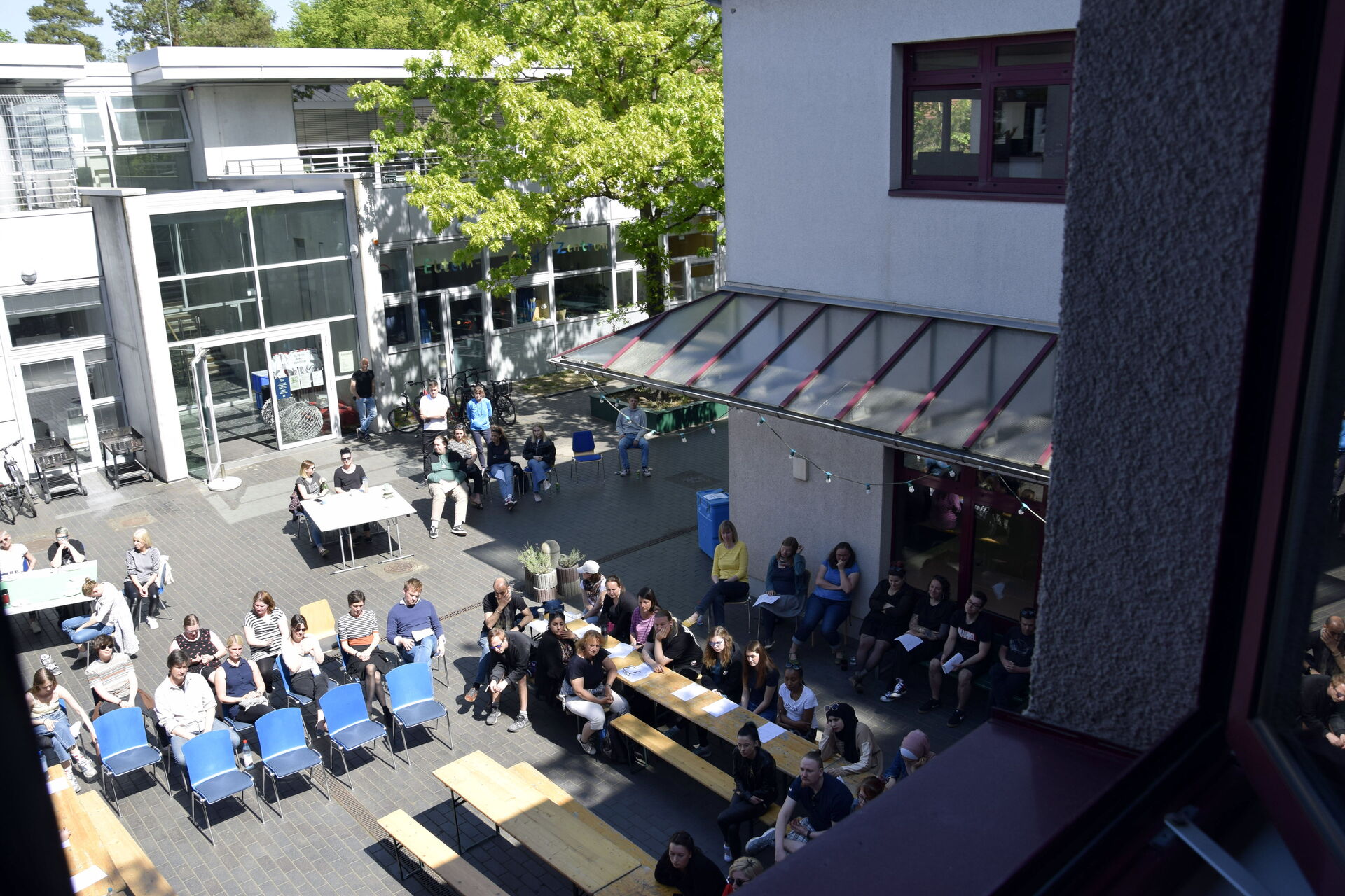 Draußen. Innenhof. Stühle und Tische an denen Menschen sitzen. Die Sonne scheint.