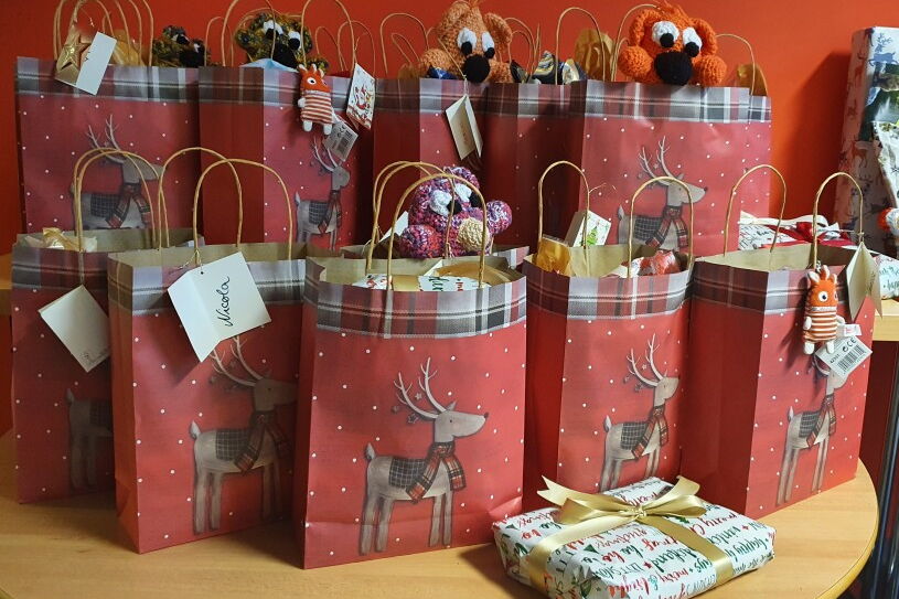 Rote Geschenktüten mit Rentier drauf, gut gefüllt stehen auf einem Tisch. Davor auf Tisch noch ein kleines Päckchen mit weißem Papier und goldenem Schleifenband.