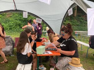 Junge Menschen sitzen unter einem Sonnensegel an einer Bierzeltgarnitur, oben am Sonnensegel ist ein Blatt mit Aufschrift: AWO Junge Plattform       angebracht.