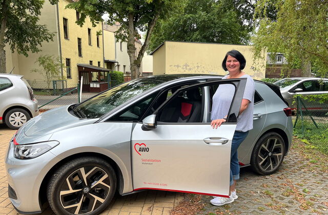 Ein silbernes VW Auto mit dem Logo der AWO Sozialstation auf der Fahrertür. Die Tür ist geöffnet und eine Frau mit schwarzen vollen Haaren, einem hellen T-Shirt und blauer dreiviertel-Jeans steht an der Fahrertür und und lächelt in die Kamera.