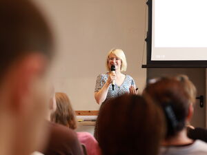 Im Vordergrund Hinterköpfe der Teilnehmenden. Vorn steht eine blonde  Frau mit Brille und Mikrofon in der Hand.