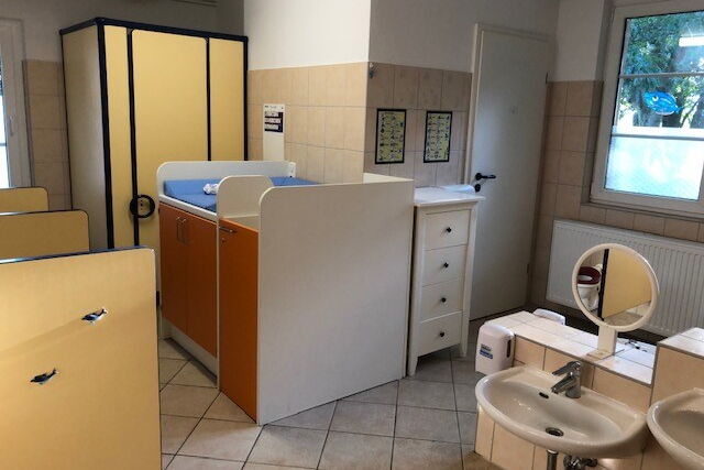 Bad mit Wickeltisch, kleinem weißen Regal und Waschbecken in verschiedenen Höhen mit Spiegel.