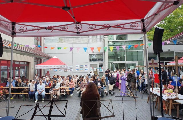 Viele Menschen auf einem Innenhof. Es gibt Biertischgarnituren, AWO Pavillons. Eine Bühne mit Lautsprechern und Notenständern. Die Menschen sehen zur Bühne.