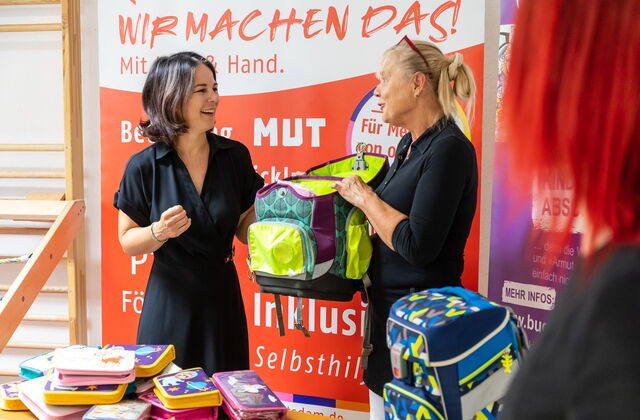 Zwei Frauen sehr fröhlich halten einen Schulranzen zwischen sich. Auf einem Tisch vor ihnen liegen mehrere bunte Federmappen für Kinder. Ein blauer Kanzen steh auf auf dem Tisch.