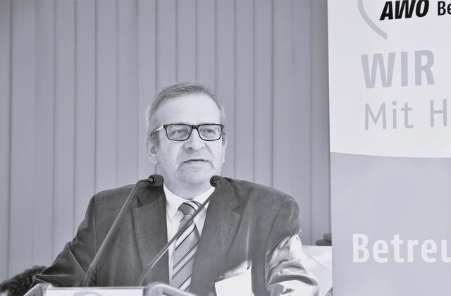 Schwarz-Weiß Foto: Mann mit Brille, Anzug, weißem Hemd in Krawatte an einem Rednerpult bei der AWO.