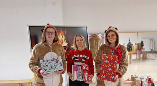 Ein heller Raum. Drei Personen stehen vor einem großen Fernseher mit Lagerfeuer-Bild. Zwei Personen tragen Rentierkostüme, Person in der Mitte mit rotem Weihnachtspulli. Alle drei halten Weihnachtsgeschenke in den Händen.