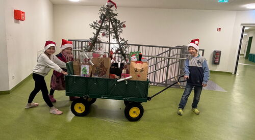 Ein Handwagen beladen mit Geschenken wir von zwei Kindern geschoben ein Junge zieht vorne am Griff. Sie sind in einem Gebäude auf einem Flur und tragen rote Weihnachtsmützen.