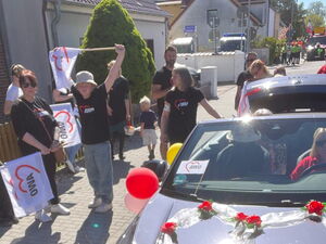 Mit Blumen und weißem AWO Fähnchen geschmücktes Auto. Links daneben Personen auch Kinder in dunklen AWO T-Shirts und weißen AWO Fahnen.