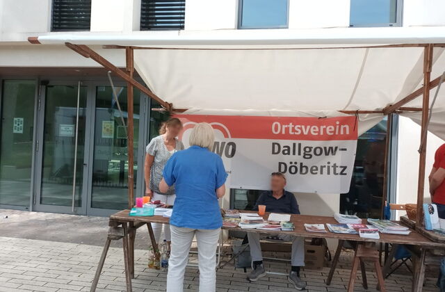 Überdachter Info-Stand des Ortsvereins. Zeitungen, Flyer, Broschüren liegen auf dem Tisch. Zwei Menschen im Stand informieren eine Person die davor steht.