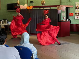 Zwei Tänzerinnen in roten Kleidern mit Kopfschmuck tanzen in einem Raum, im Vordergrund sitzen Zuschauer.