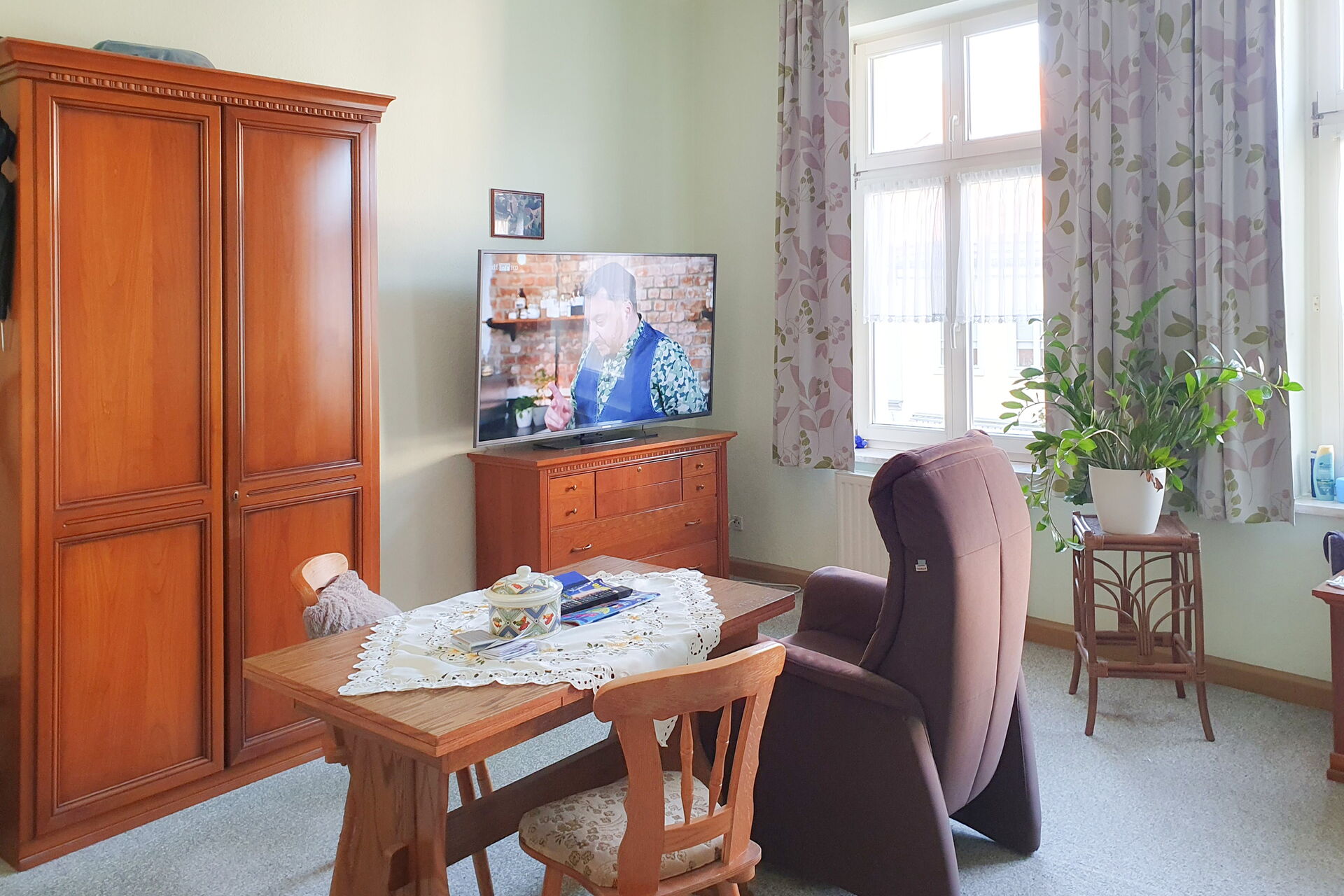 Raum mit Holzschrank und Fernsehschränkchen an der Wand. Fernseher ist an. Ein Fernsehsessel steht im Raum, daneben ein Tisch mit zwei Stühlen.