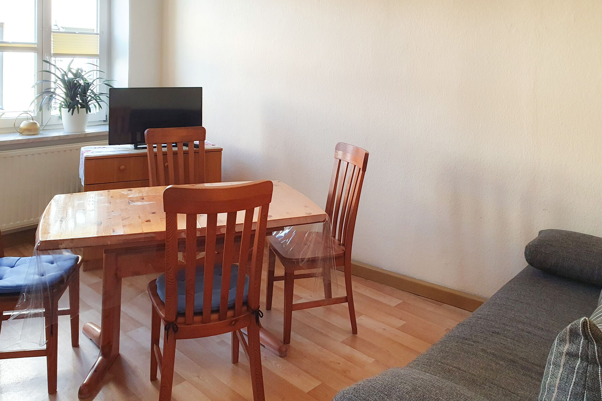 Zimmer mit grauer Couch in der rechten Bildecke. Davor ein brauner Holztisch mit vier Stühlen und der Couch gegenüber ein Fernseher auf einem kleinen Schrank/Kommode.