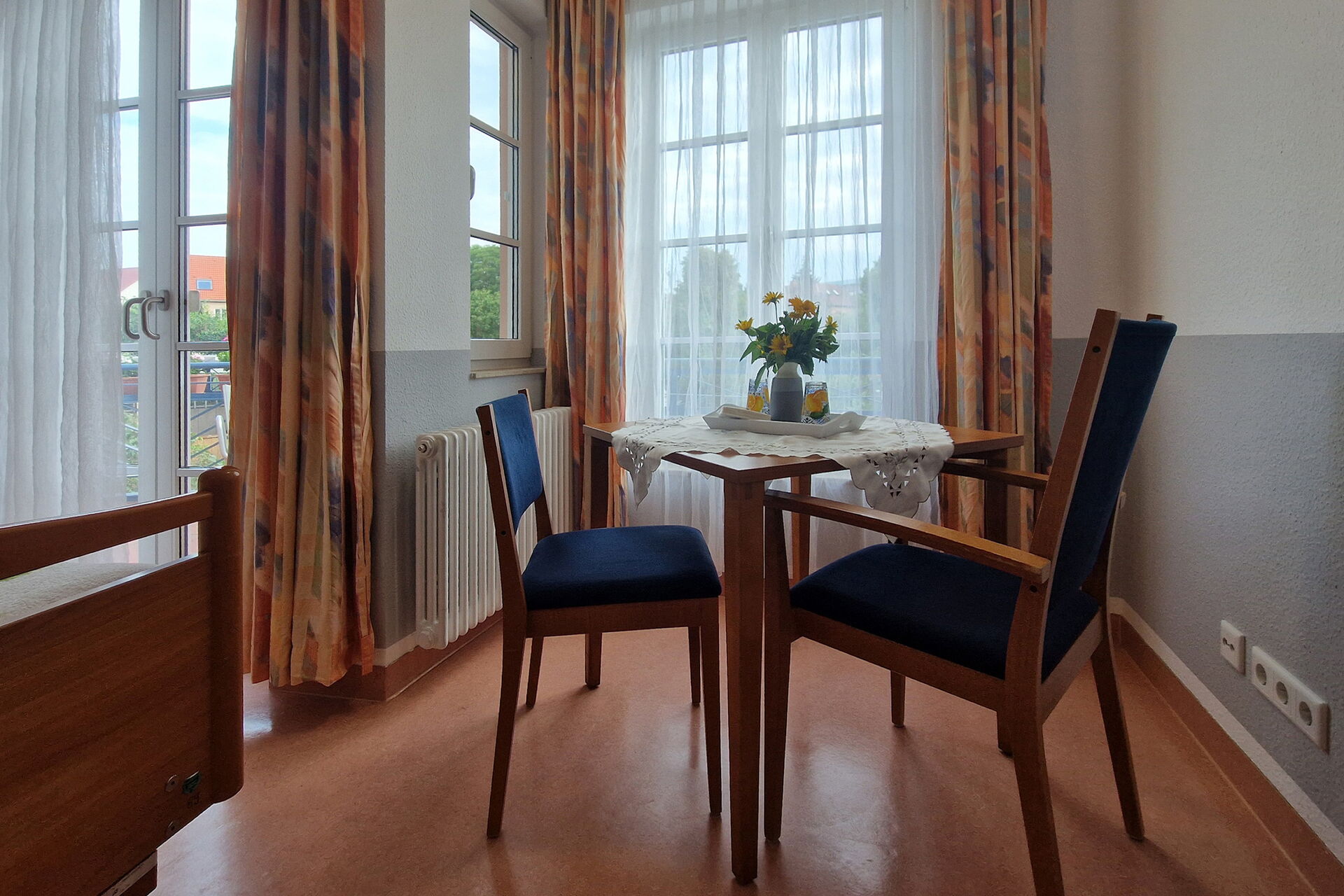 Zimmer, zwei Stühle blau gepolstert und kleiner Tisch vor Fenstern.