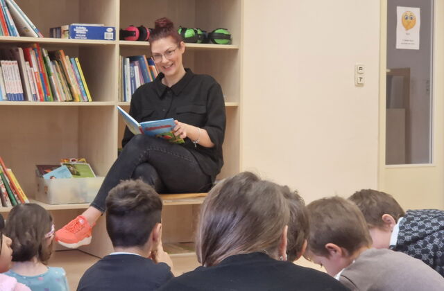 Eine Frau sitzt mit einem Buch in der Hand in einem Raum mit Regalen. Sie lächelt die Kinder, die vor ihr auf dem Boden sitzen an.