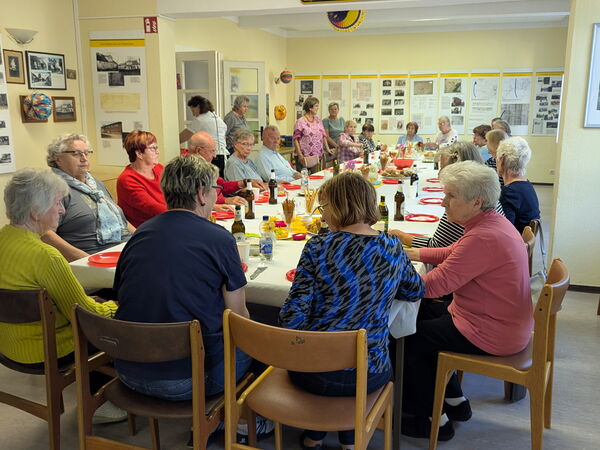 Großer Raum, breite, lange gedeckte Tafel. Viele Menschen sitzen mit Getränken und Snacks versorgt am Tisch.