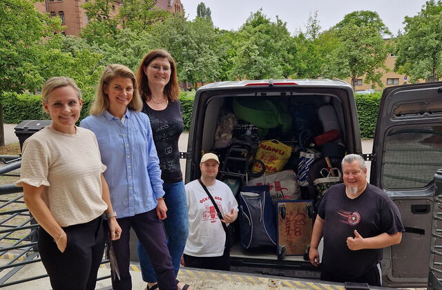 3 Frauen stehen im Vordergrund auf einer Erhöhung. Dahinter und unten auf Straße ein vollgepackter Transporter von hinten, beide Türen geöffnet. Zwei Männer stehen vor dem Laderaum machen ein Daumen-Hoch.