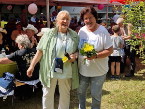 Zwei Frauen stehen in der Sonne auf einer Wiese und lächeln in die Kamera. Dahinter AWO Pavillons und Menschen an Tischen.