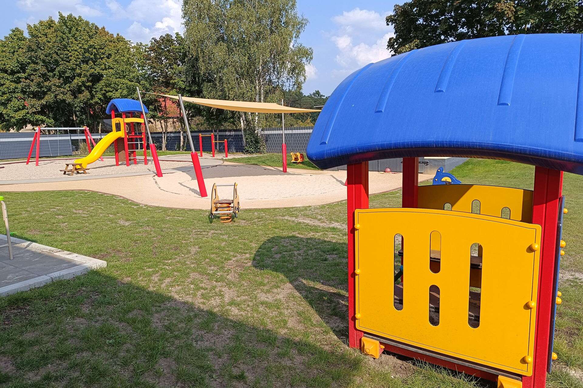 Spielplatz, Sandkasten, Schaukel, Klettergerüst mit Rutsche. Wiese Bäume, die Sonne scheint.