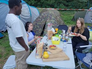 Frühstück auf dem Campingplatz. Zwei weiße Fraue sitzen auf Campingstühlen an einem Klapptisch mit Toast, Zitronen, Wasser Flasche. Ein schwarzer Mann steht am Tisch, Hände in den Hosentaschen. Zelte stehen ringsum.