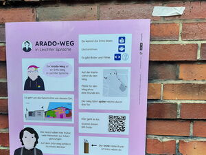 Ein hellila gerahmtes Schild ist an einer Backsteinwand befestigt. Auf dem Schild steht ganz oben "Arado-Weg in Leichter Sprache". Darunter sind weiße Kästchen mit Symbolen und Schrift, in denen der Wegabschnitt in Leichter Sprache erklärt wird.