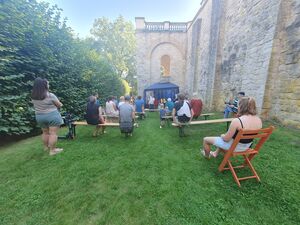 Draußen, schönes Wetter. Eine Wiese mit Bierbänken. Menschen sitzen dort und sehen sich etwas an. Vorn ist ein blauer Pavillon wo etwas aufgeführt wird. Die Mauern des Belvedere und hohe grüne Ecken Rahmen das Ganze ein.