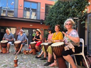 Draußen, 6 Menschen sitzen auf Stühlen und haben Djembe / Trommeln zwischen den Beinen und musizieren gemeinsam.