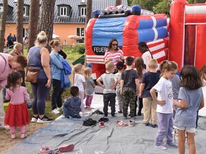 Eine Feuerwehr-Hüpfburg zum raufklettern und runterrutschen. Einige Kinder stehen davor und haben ihre Schuhe ausgezogen, Eine Frau mit Sonnenbrille und braunen langen Haaren erklärt etwas. Mütter und Großeltern stehen bei den Kindern.