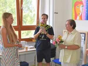 Beide Künstler stehen nebenienander und halten kleine Blumensträuße im Wasserglas in der Hand. Jüngerer Mann in blauem T-Shirt und kurze Hose. Ältere Frau, kurze graue Haare und Brille mit heller Bluse. Freu mit langen blonden Haaren steht ihnen gegenüber