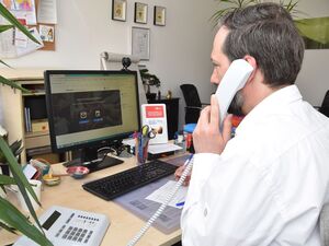 Ein Mann in weißem Hemd sitzt in einem NBüro am Schreibtisch, vor ihhm der Monitor, Tastatur unnd ein Notizblock, wo er etwas notiert, er hat den Telefonhörer am Ohr.