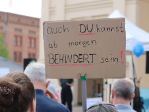 Menschen von hinten. Eine Person hält Protestschild hoch. Darauf steht: auch du kannst ab morgen behindert sein!