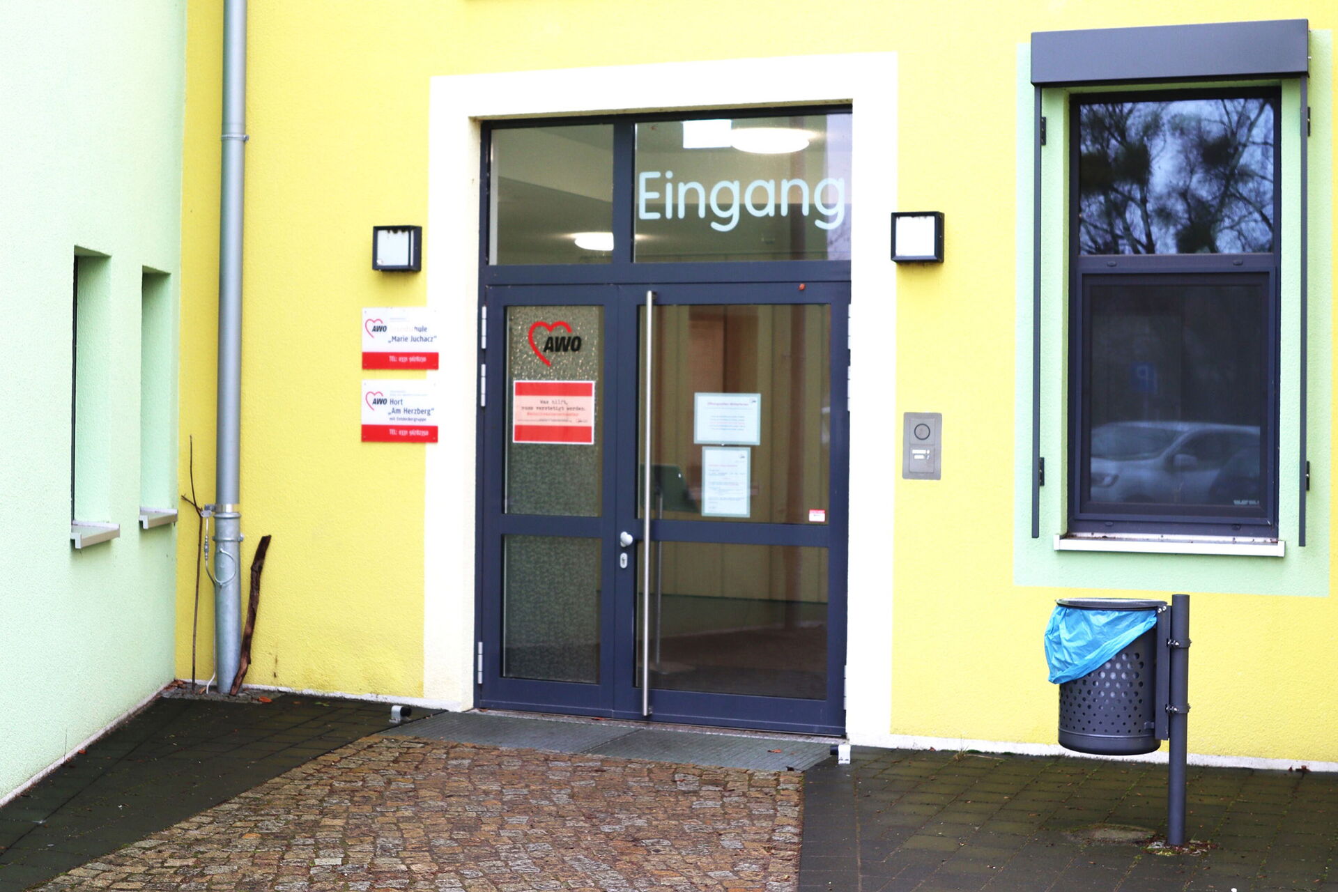 Eingangstür ist geschlossen. AWO Logo an der Glastür. Das Gebäude ist gelb. Im Fenster spiegelt sich ein Auto. Ein Mülleiner ist Eingangsbereich.