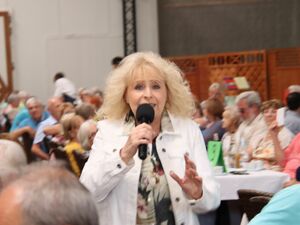 Blonde schlanke Frau mit lockigen Haaren und Ponny in der Stirn. Sie hat ein Mikro in der Hand. Sie trägt eine helle leichte Jacke und darunter ein Bluse mit Natur/Blumen Muster. Sie steht in Mitten von Menschen die an Tischen sitzen.