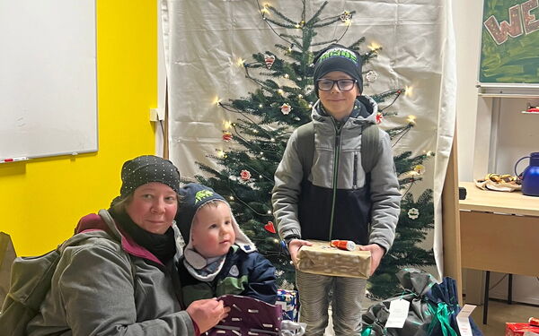 Geschenkeübergabe. Klassenraum. Frau hockt bei einem kleinen Kind, das Kind hat ein großes Geschenk. Vor einem kleinen Weihnachtsbaum, steht ein Junge auch mit Geschenk in den Händen.